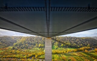 A picture of the underside of a steel bridge spanning a valley full of green trees, fields, and pastures.