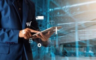 Angle on a man in a blue blazer standing in a steel shipping facility typing on his automated transaction processing device. A graphic design overlays the image, consisting of various icons representing Steel Supply Chain Management.