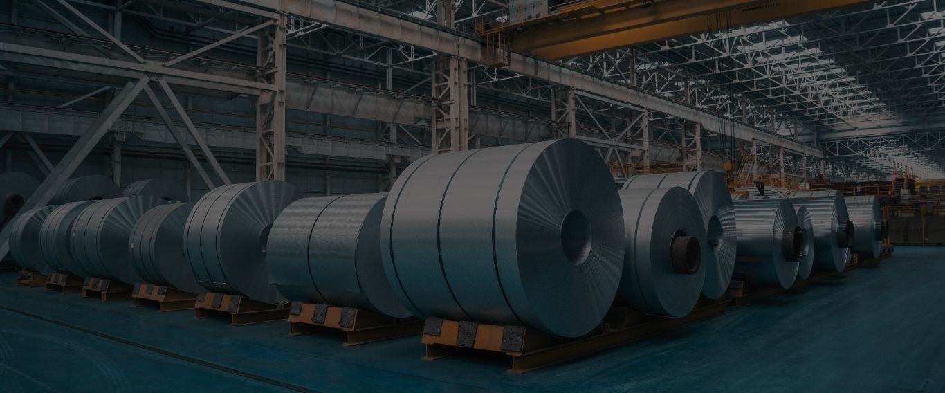 Rows of massive rolled steel coils stored on platforms inside a spacious industrial warehouse with high ceilings and overhead cranes.