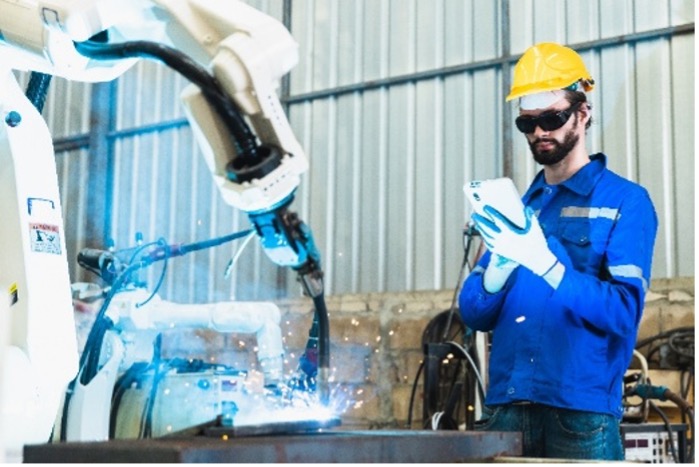 An individual in a yellow hard hat, blue protective clothing, and gloves is operating a tablet device next to a robotic arm that is welding, producing sparks. The setting is an industrial or manufacturing environment with metal walls and various equipment in the background.