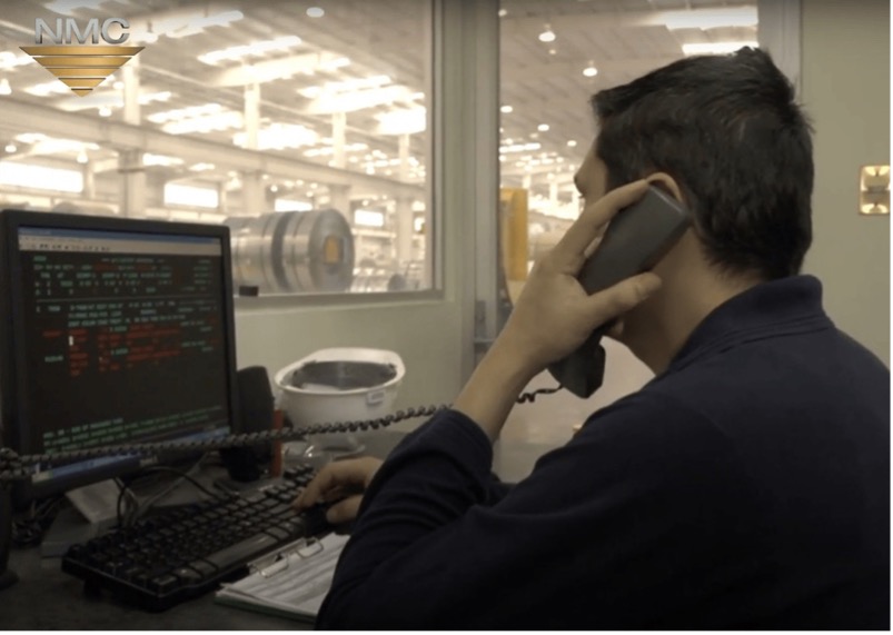 An industrial operator at a control station, analyzing data on a large monitor with multicolored code or system outputs. The setting emphasizes real-time oversight and digital integration in metal supply chains, with the NMC logo reinforcing the industry context.